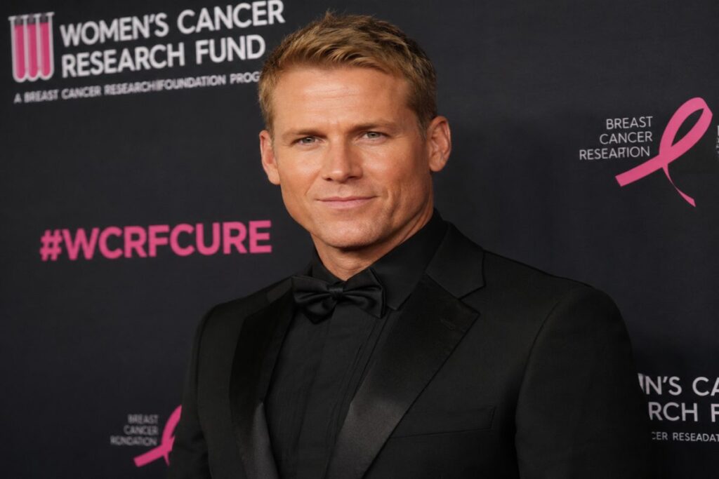 Jason Schanne posing in a black tuxedo at a formal red carpet event, standing confidently in front of an event-branded backdrop.